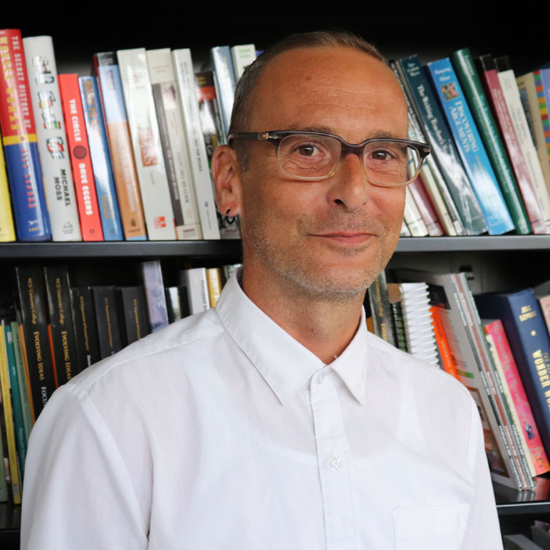A man with dark-rimmed glasses wearing a white dress shirt standing in front of a large bookcase