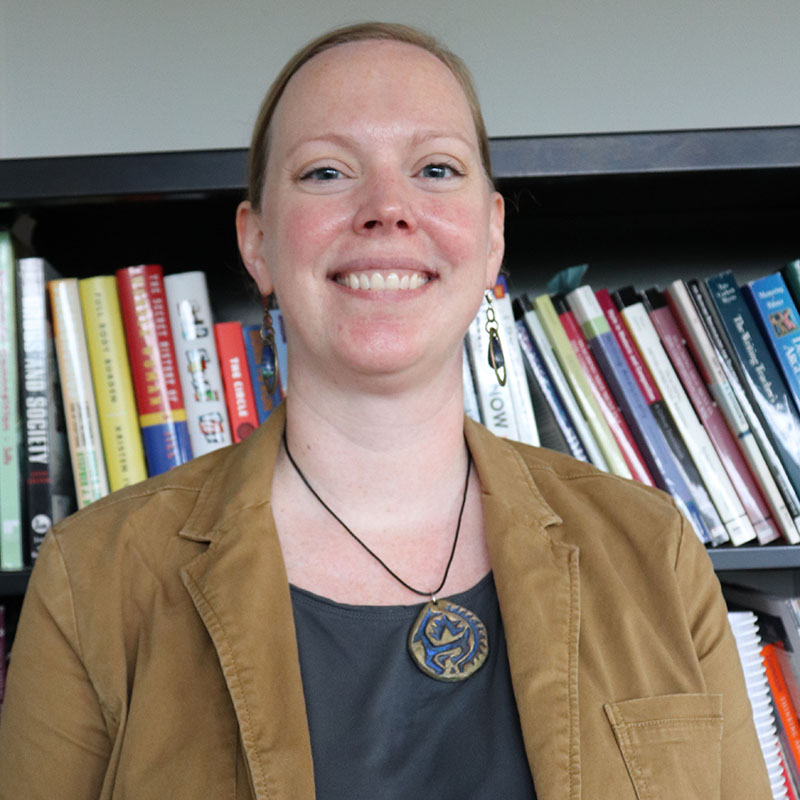 A woman wearing a bown earth tone jacket with a gray shirt and necklace