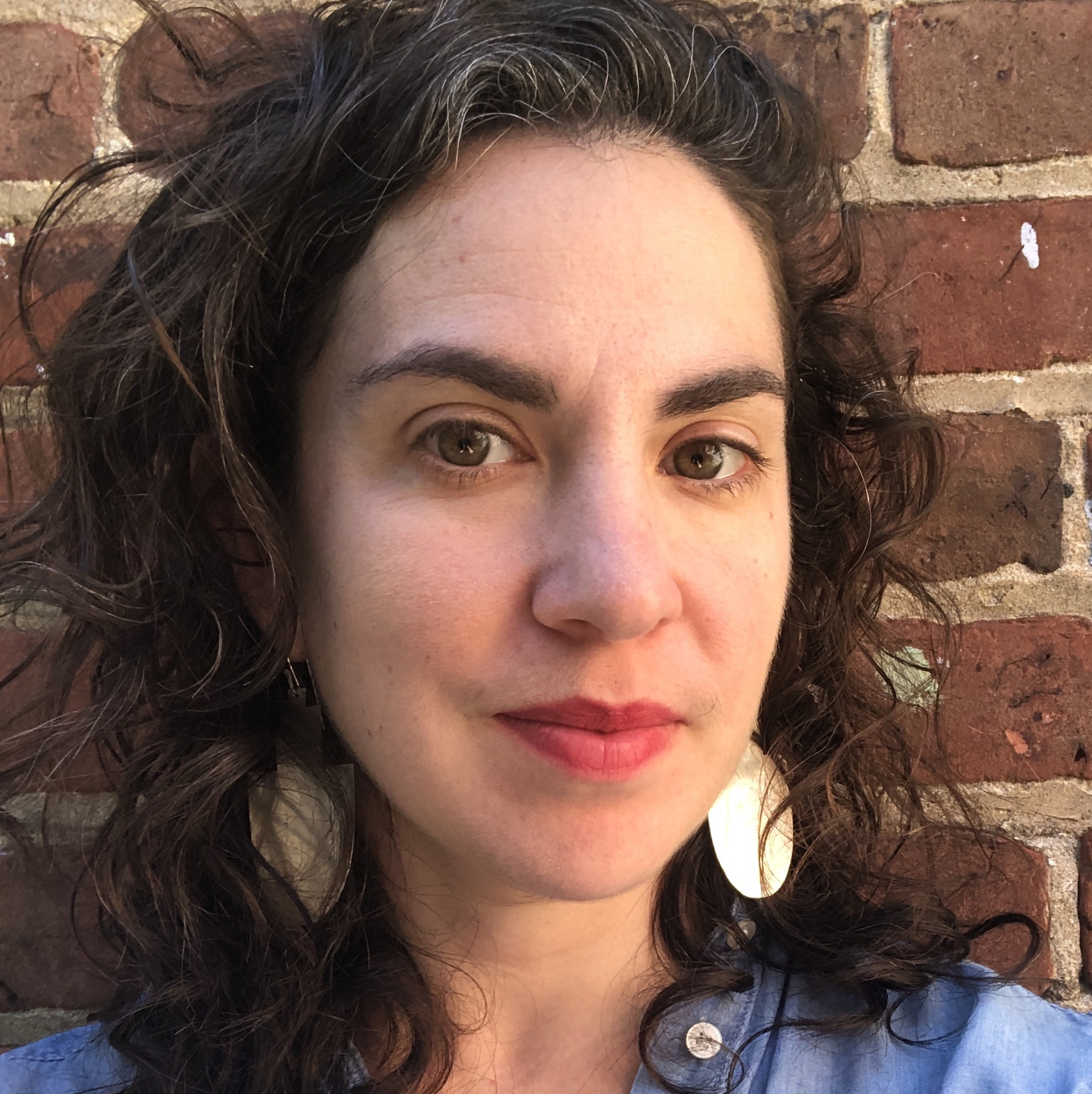 A woman wearing a denim blue shirt with earrings stadning in front of a brick wall