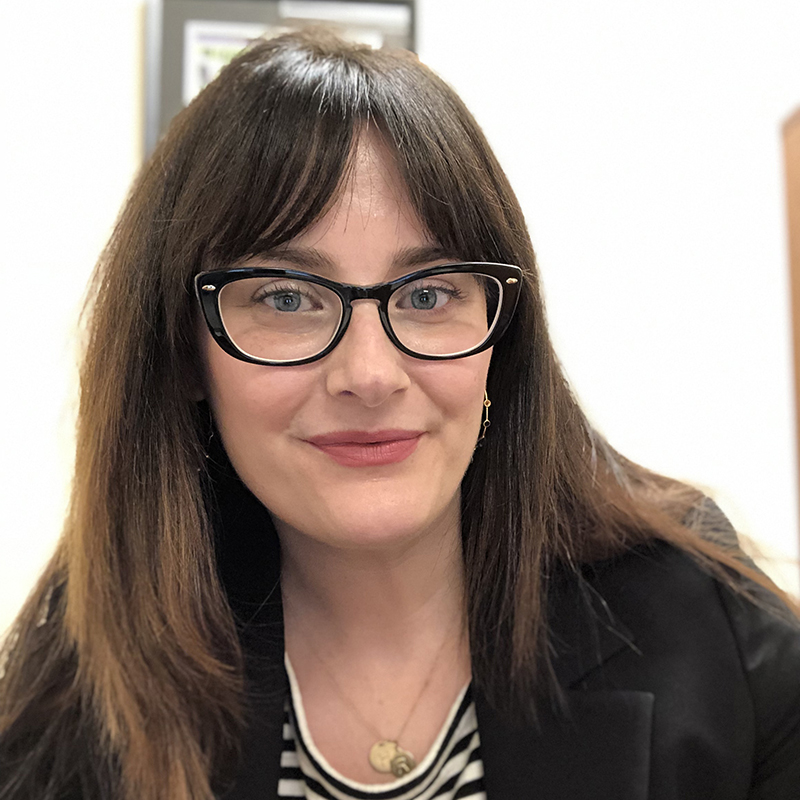 A woman wearing black rimmed glasses with a black blazer and light colored blouse