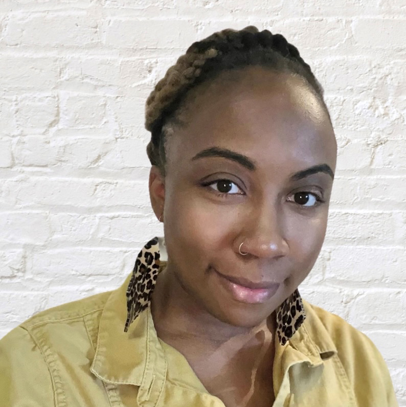 A woman wearing a gold top with earrings standing in front of a painted white brick wall