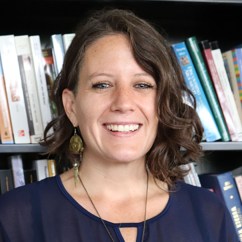 A woman standing in front of a bookcase wearing a navy blue blouse and necklace