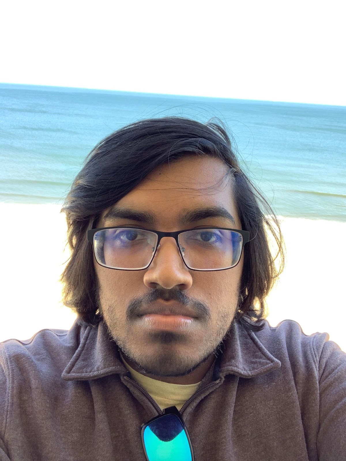 A man with dark black hair wearing glasses standing in front of the ocean